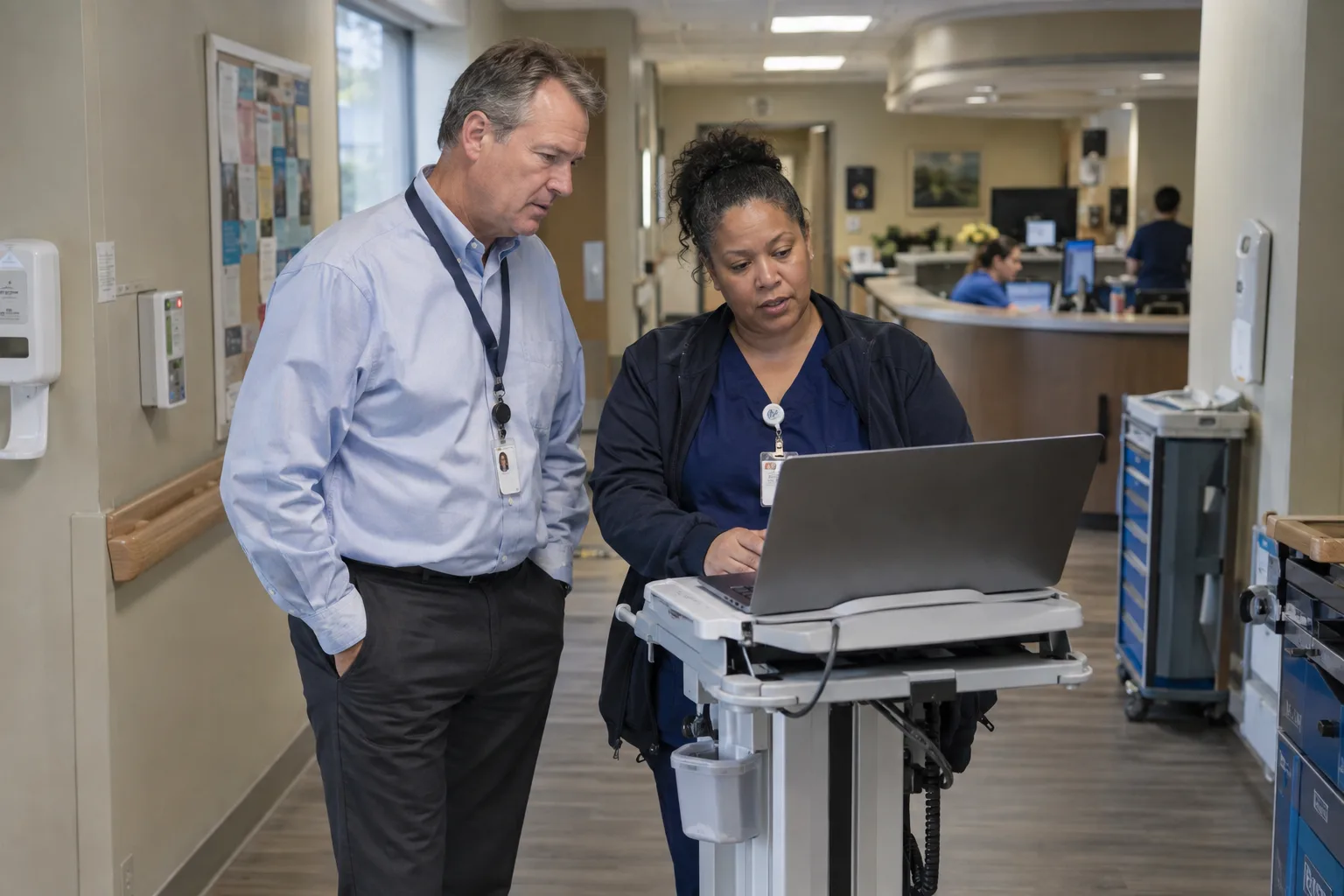An administrator and a director of nursing review a laptop together in a nursing unit hallway.